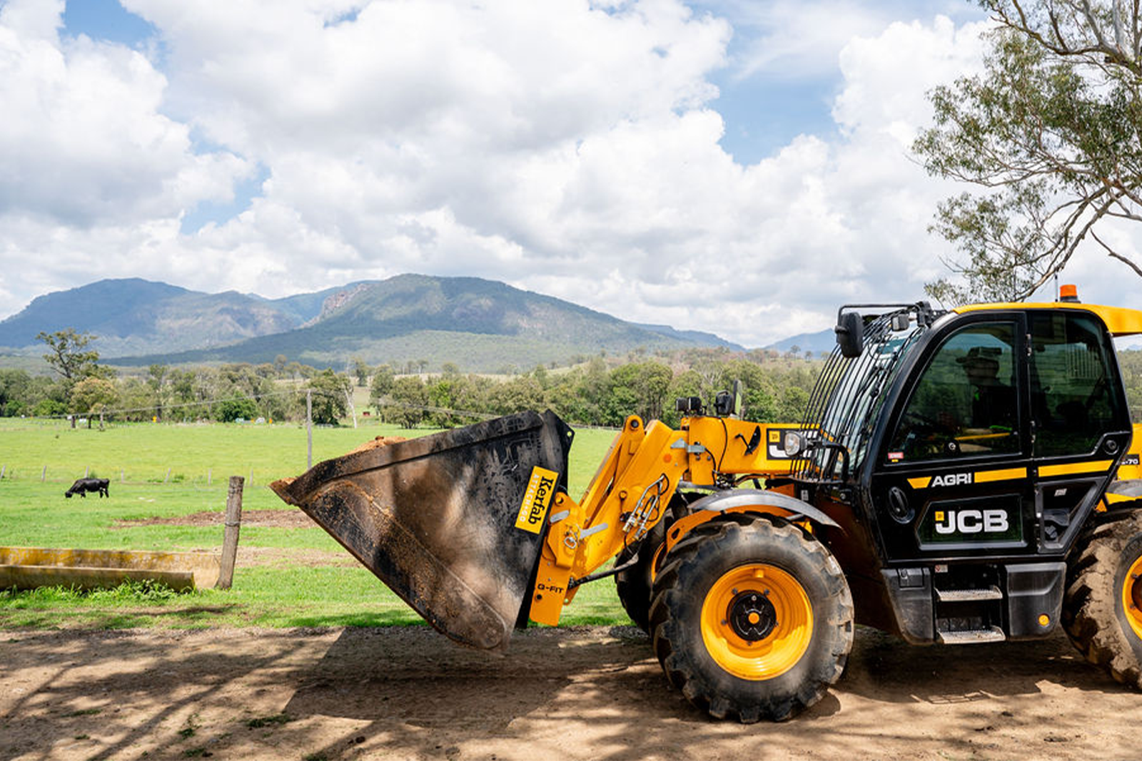 JCB telehandler fitted with a Kerfab Attach + Go bucket working on a farm; Kerfab logo visible, green paddocks and mountains in the background.