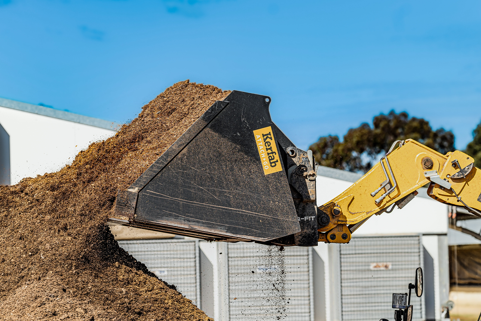 Kerfab Civil Construction Attachments — loader bucket moving a large pile of soil; black bucket with yellow Kerfab logo raised against blue sky, industrial bins in background.