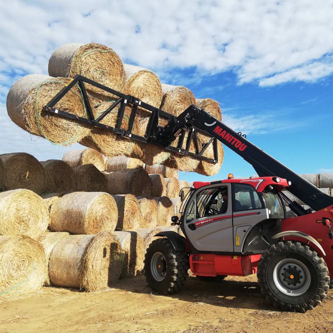 Telehandler using a Kerfab multi-bale grab to lift and stack round hay bales; red machine working in a farm yard under blue sky.