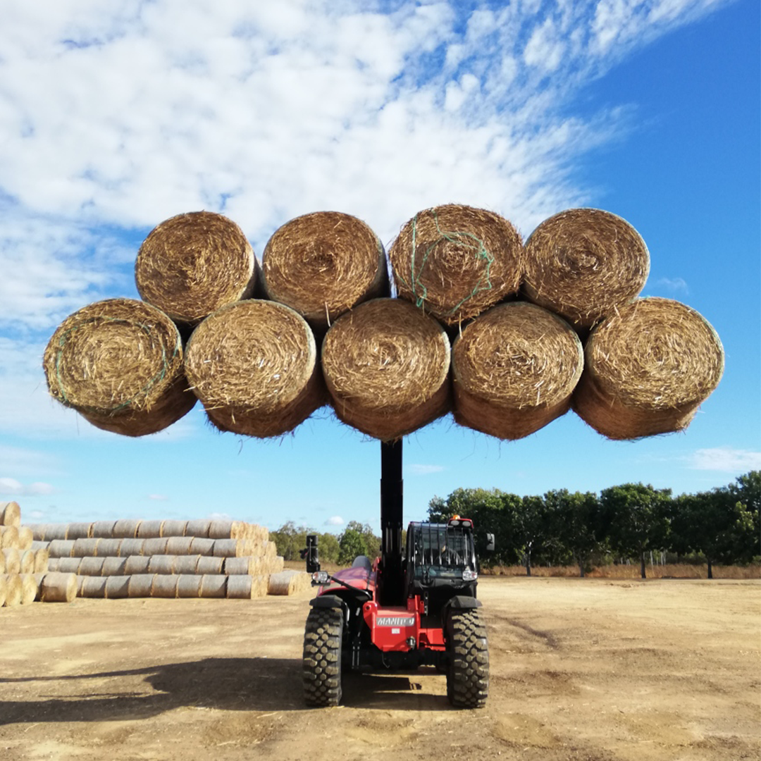 Telehandler lifting a large stack of round hay bales using a bale spike/fork — showcasing Kerfab hay-handling attachments in a farm yard under blue sky.