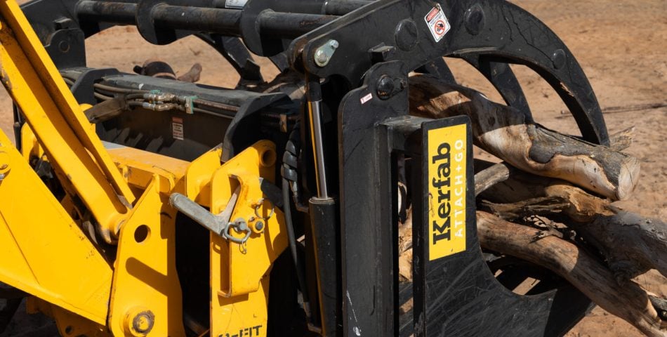 Close-up of Kerfab ATTACH+GO Stone Fork Grapple on a JCB telehandler gripping logs, showing hydraulic clamp and quick-hitch in a sandy paddock.